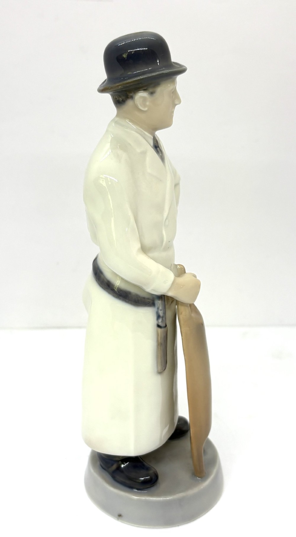 A glazed porcelain figurine depicting a butcher wearing a bowler hat, double-breasted coat, and apron standing beside a brown trough. View II