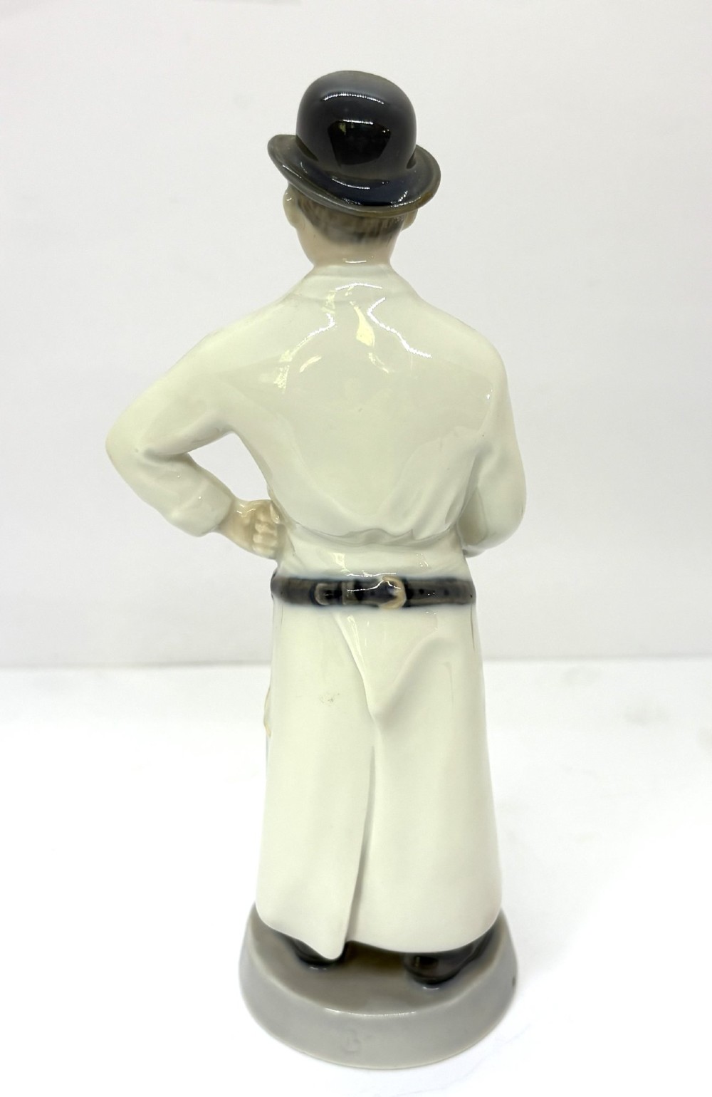 A glazed porcelain figurine depicting a butcher wearing a bowler hat, double-breasted coat, and apron standing beside a brown trough. View III
