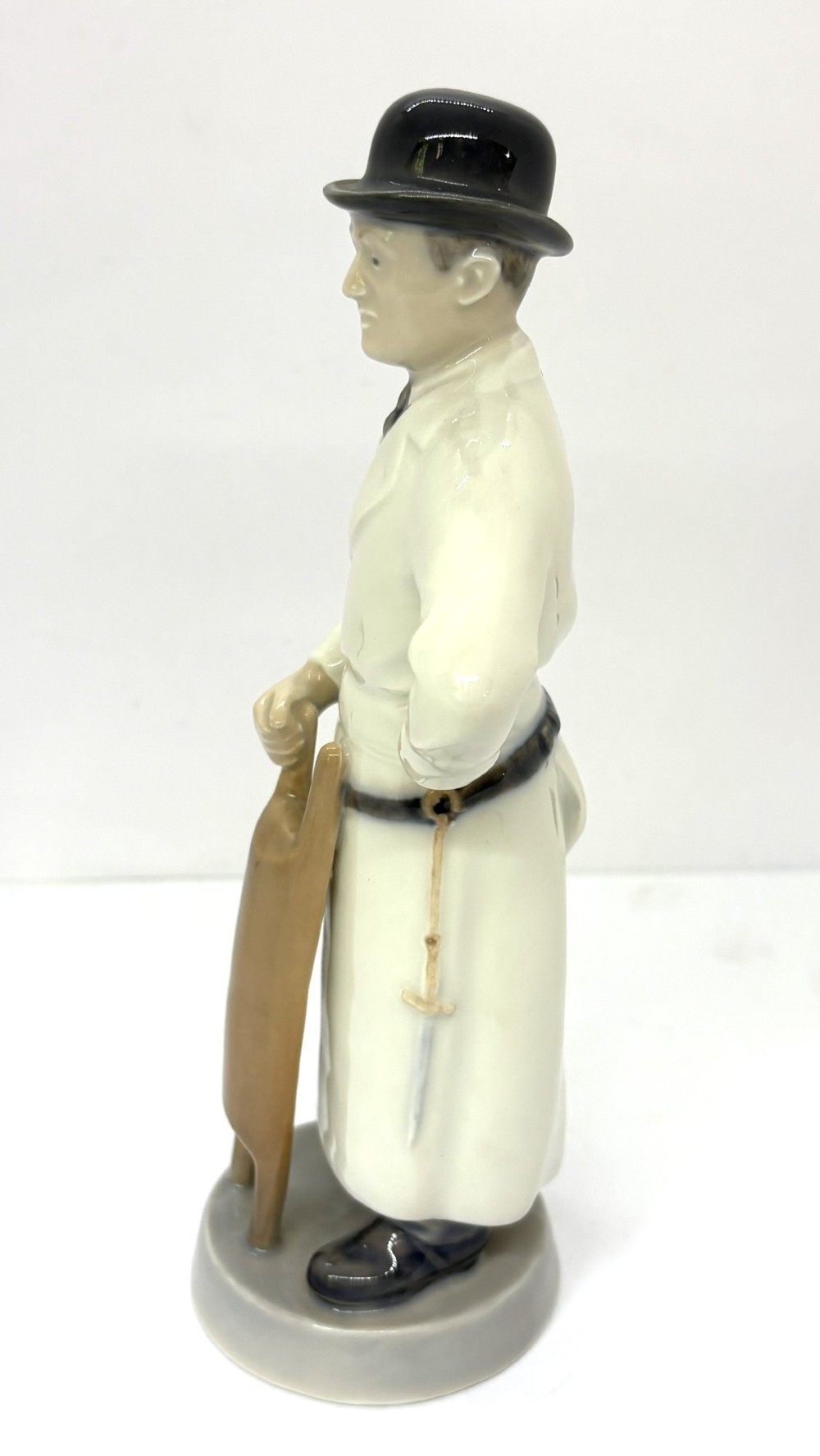 A glazed porcelain figurine depicting a butcher wearing a bowler hat, double-breasted coat, and apron standing beside a brown trough. View IV