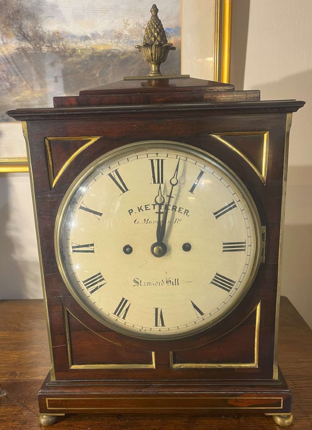A Regency mahogany bracket clock featuring brass corner inlay, a top pineapple finial, brass ball feet, and a cream painted dial signed P. Ketterer Stamford Hill.
