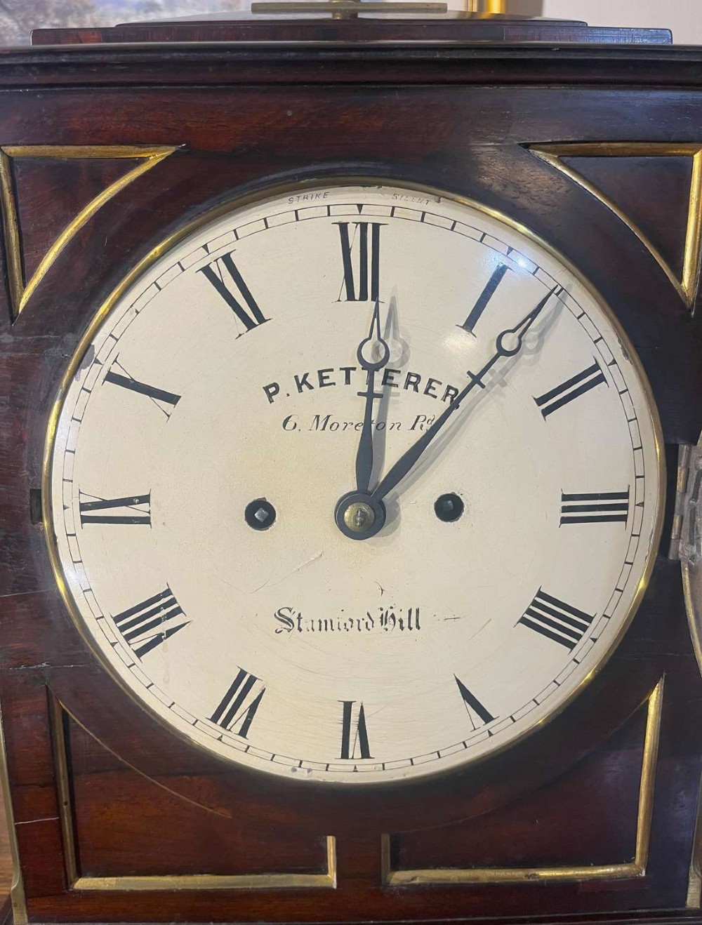 A Regency mahogany bracket clock featuring brass corner inlay, a top pineapple finial, brass ball feet, and a cream painted dial signed P. Ketterer Stamford Hill. View II