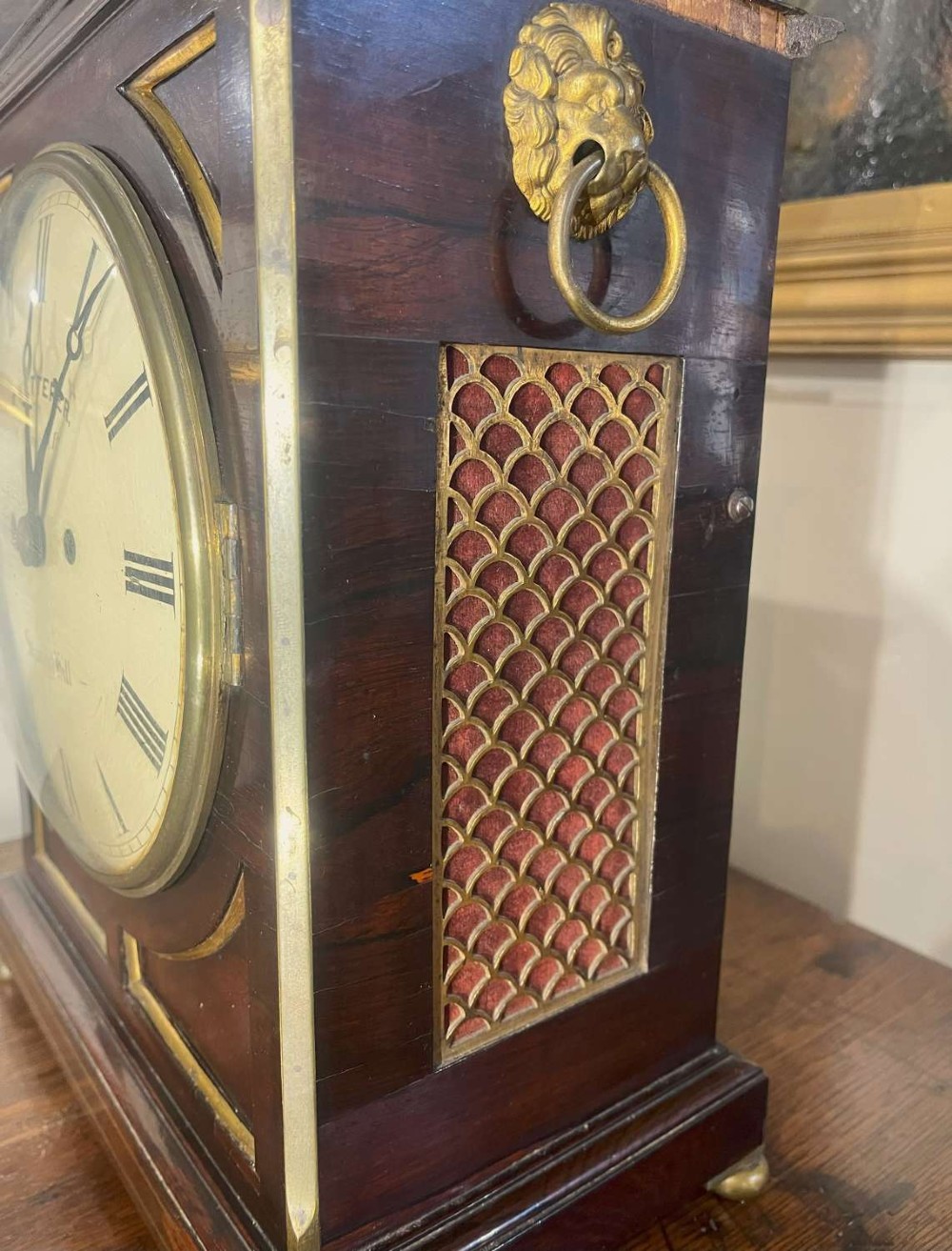 A Regency mahogany bracket clock featuring brass corner inlay, a top pineapple finial, brass ball feet, and a cream painted dial signed P. Ketterer Stamford Hill. View V