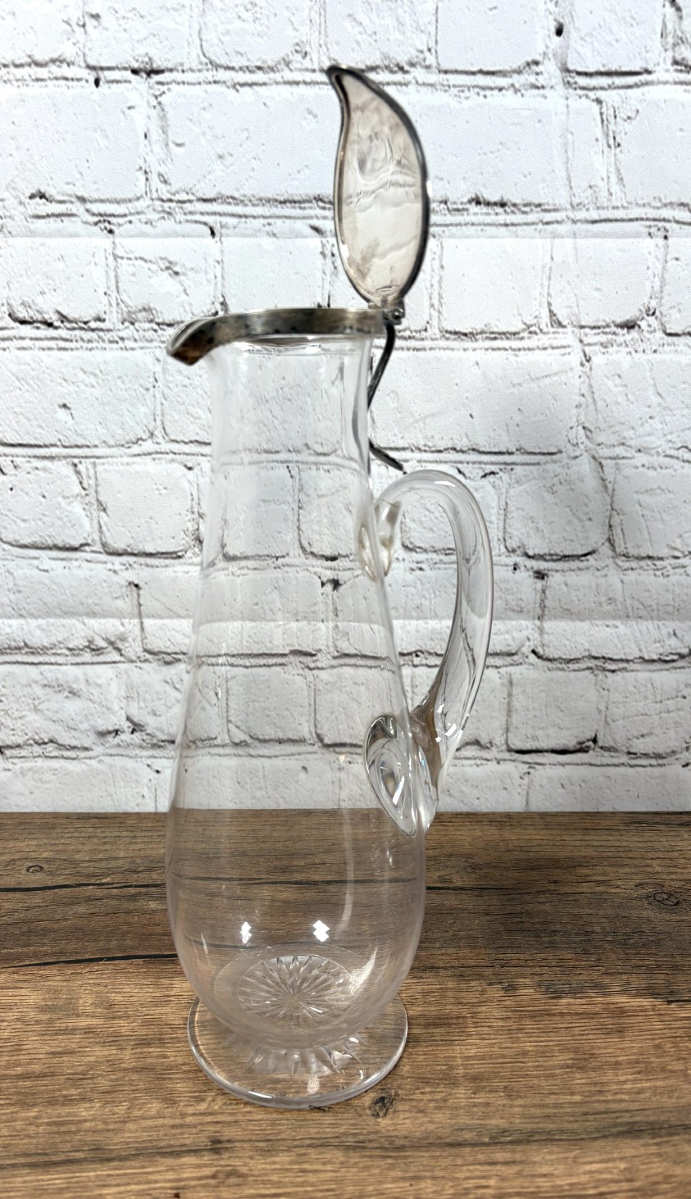 A clear glass claret jug featuring a starburst cut base, a loop glass handle, and an open hinged silver lid.