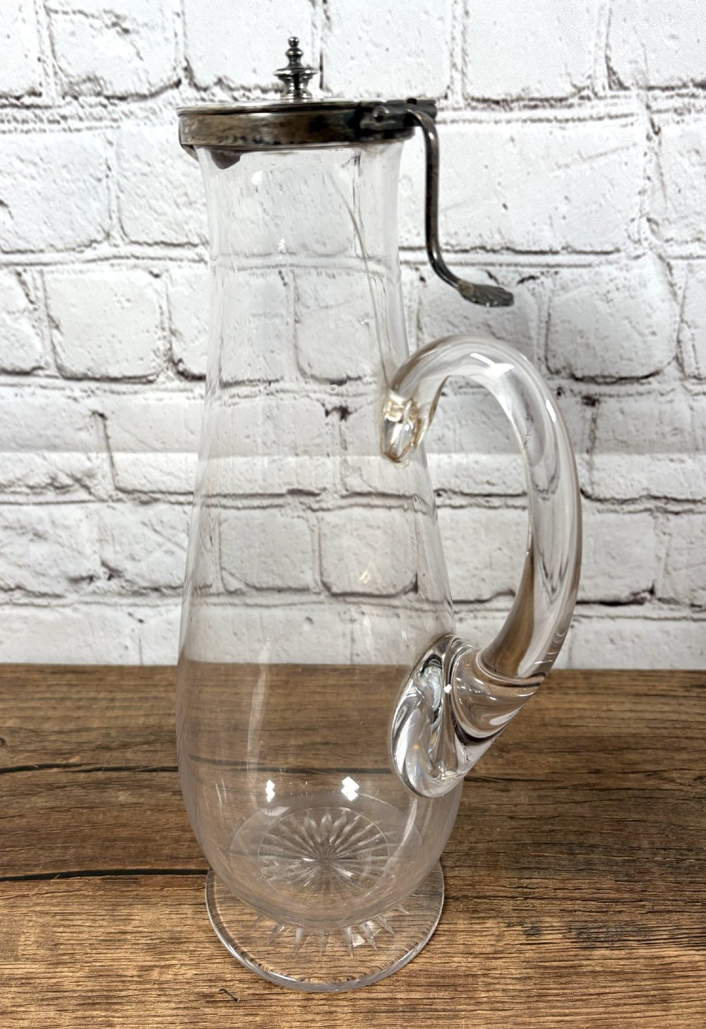 A clear glass claret jug featuring a starburst cut base, a loop glass handle, and an open hinged silver lid. View III