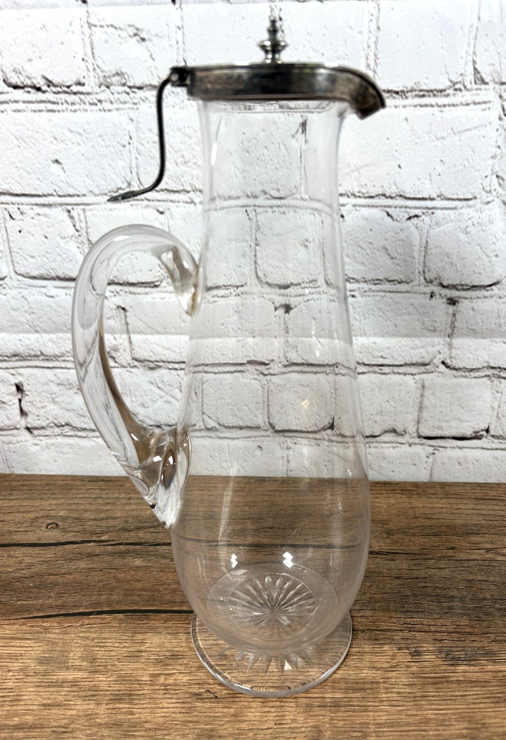 A clear glass claret jug featuring a starburst cut base, a loop glass handle, and an open hinged silver lid. View IV