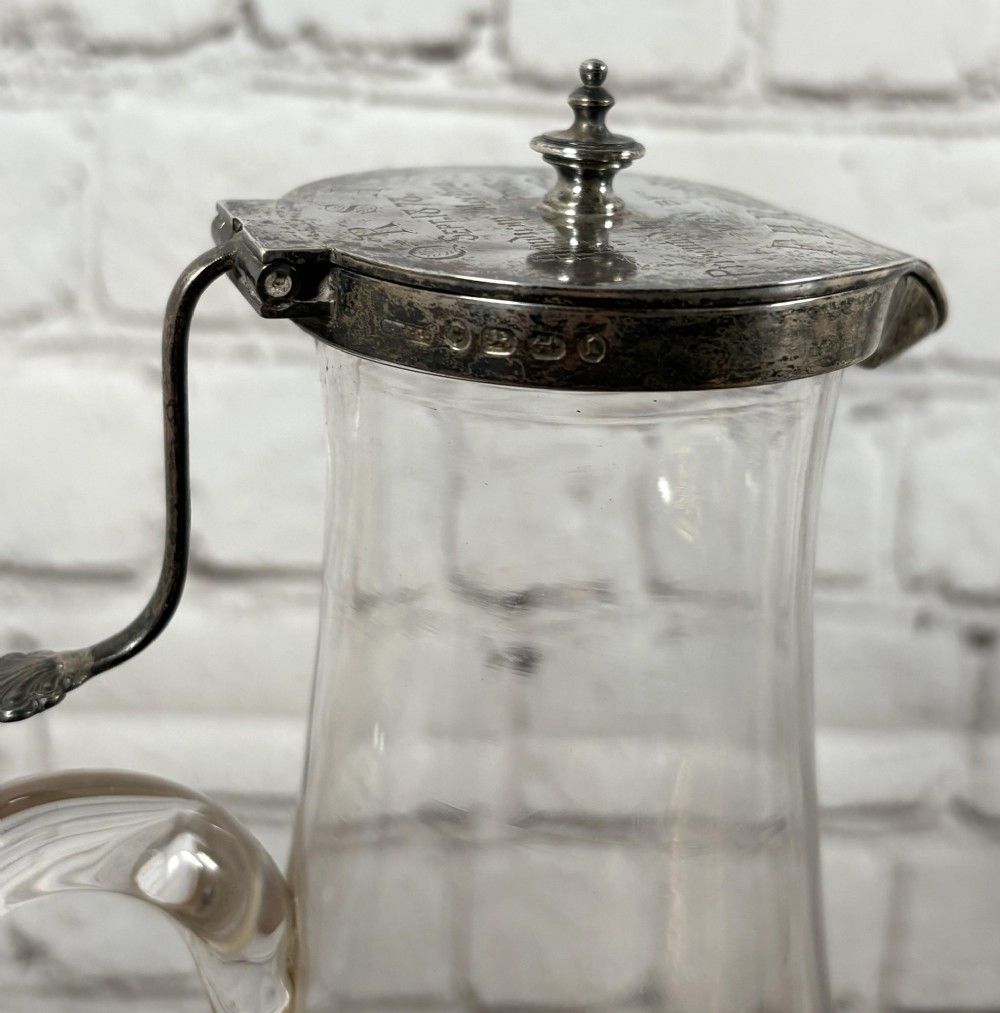 A clear glass claret jug featuring a starburst cut base, a loop glass handle, and an open hinged silver lid. View VI