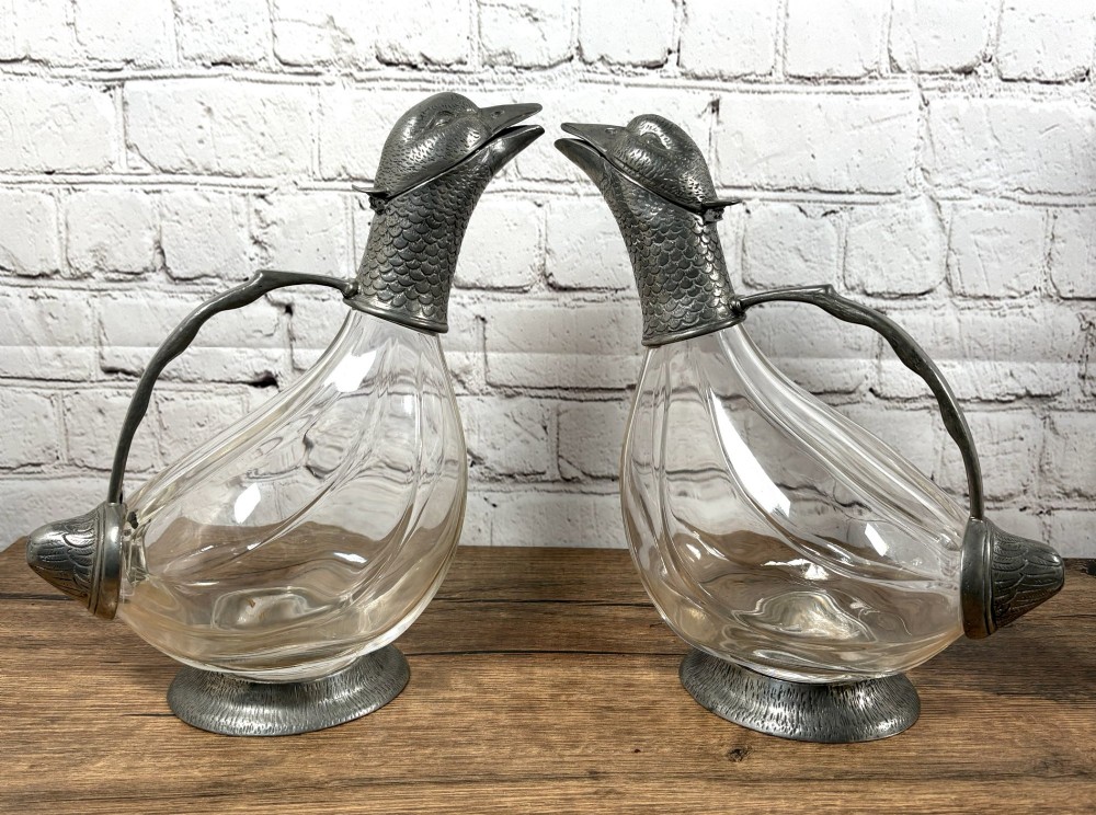 A pair of clear glass claret jugs shaped like ducks with pewter heads, handles, tail caps, and circular bases.