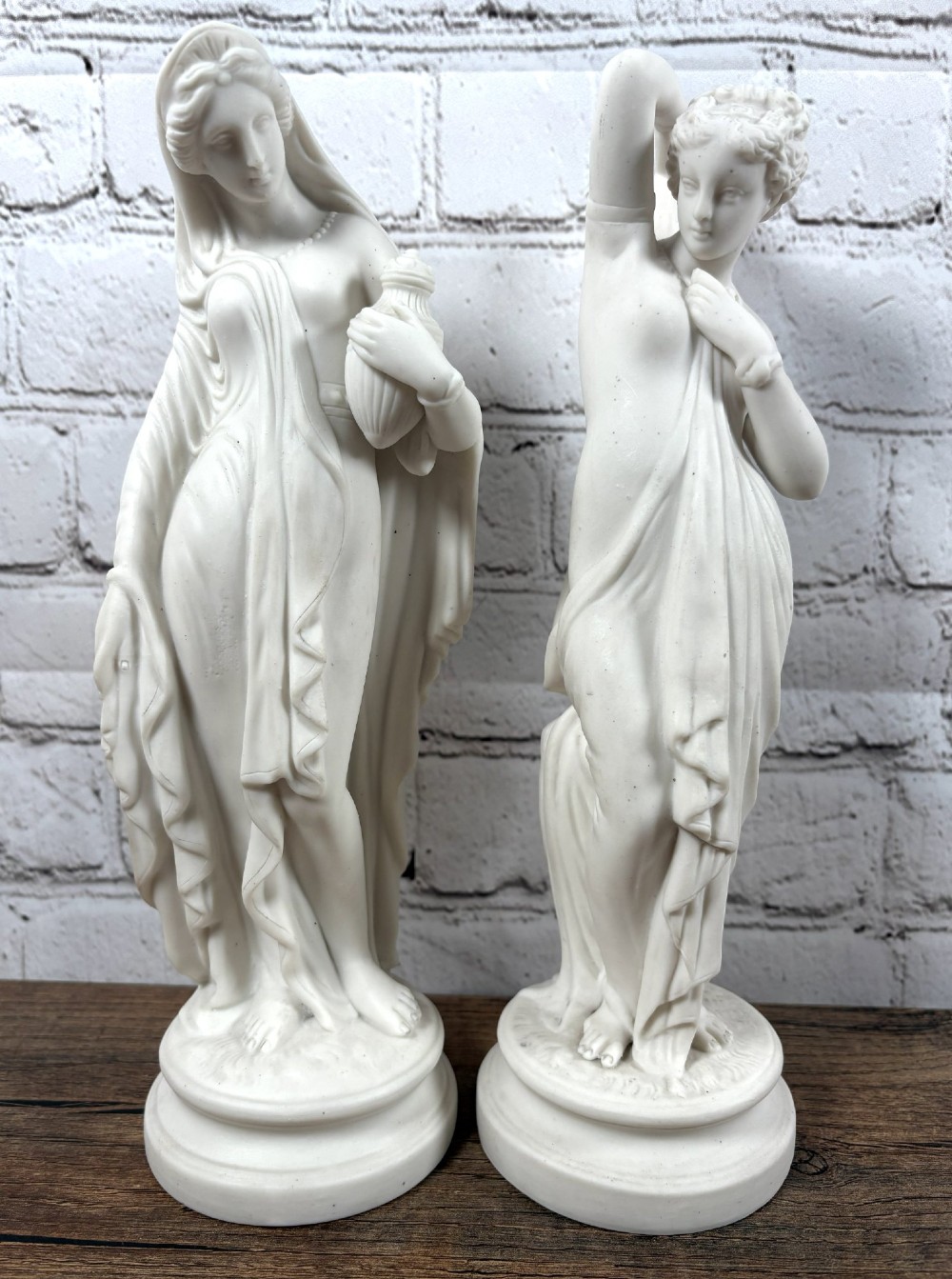 A pair of 19th-century unglazed white Parian ware porcelain figures depicting classical maidens in draped garments, standing on circular integrally moulded plinths.