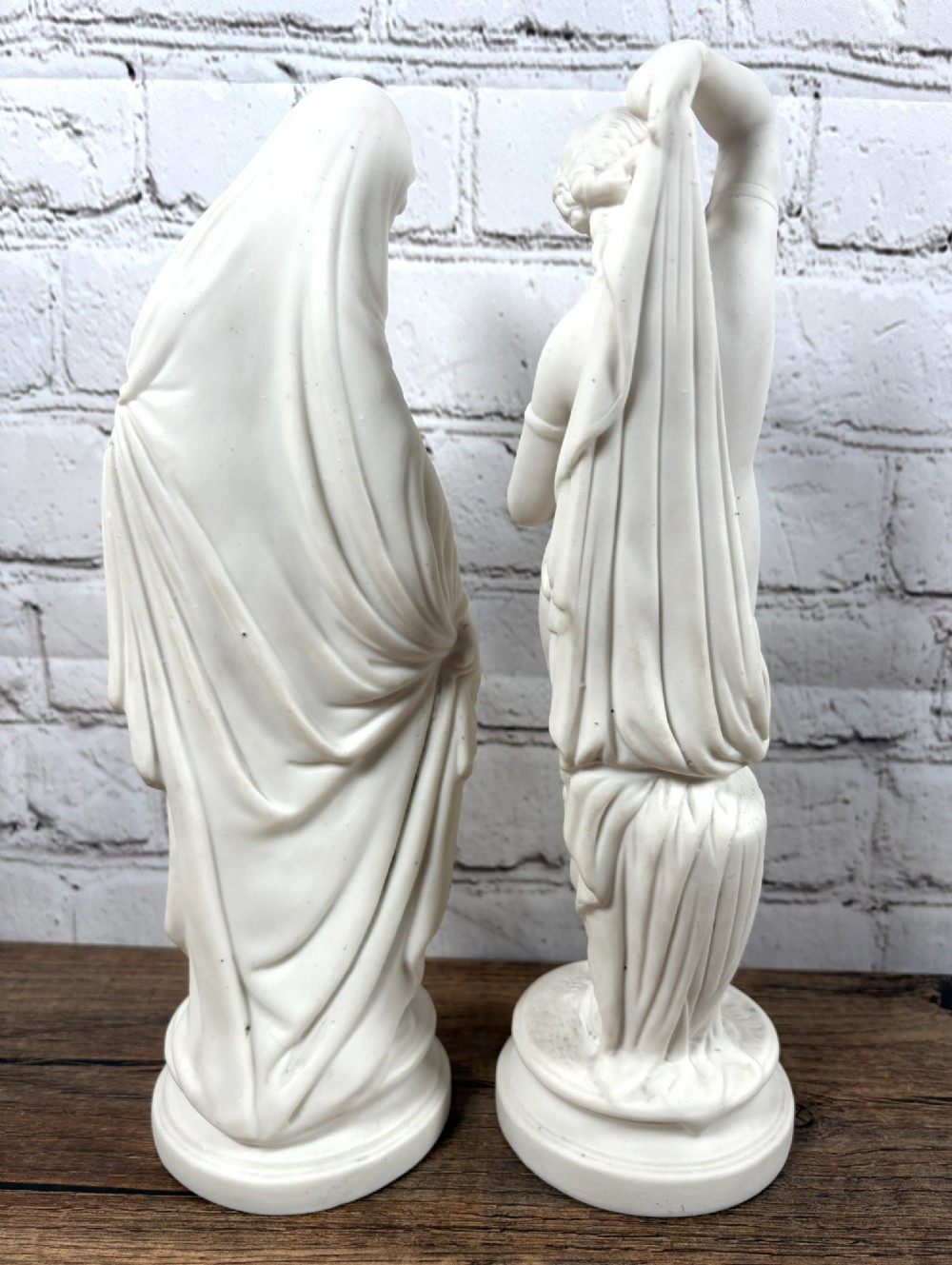 A pair of 19th-century unglazed white Parian ware porcelain figures depicting classical maidens in draped garments, standing on circular integrally moulded plinths. View IV