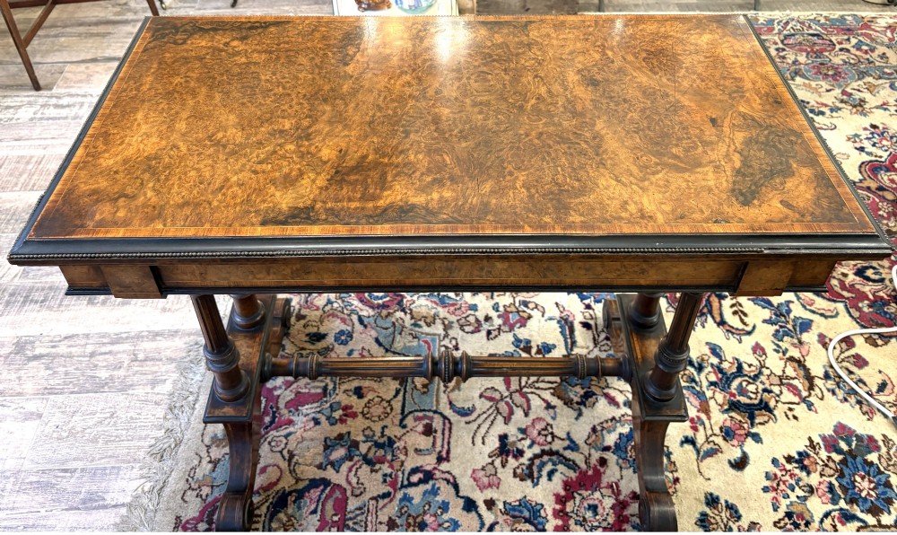A Victorian burr walnut card table featuring ebonised bead moulding, crossbanded veneer top, twin turned pillar supports, and a turned stretcher base.