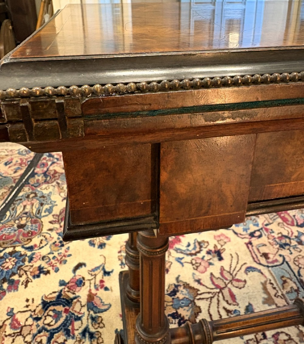A Victorian burr walnut card table featuring ebonised bead moulding, crossbanded veneer top, twin turned pillar supports, and a turned stretcher base. View VII