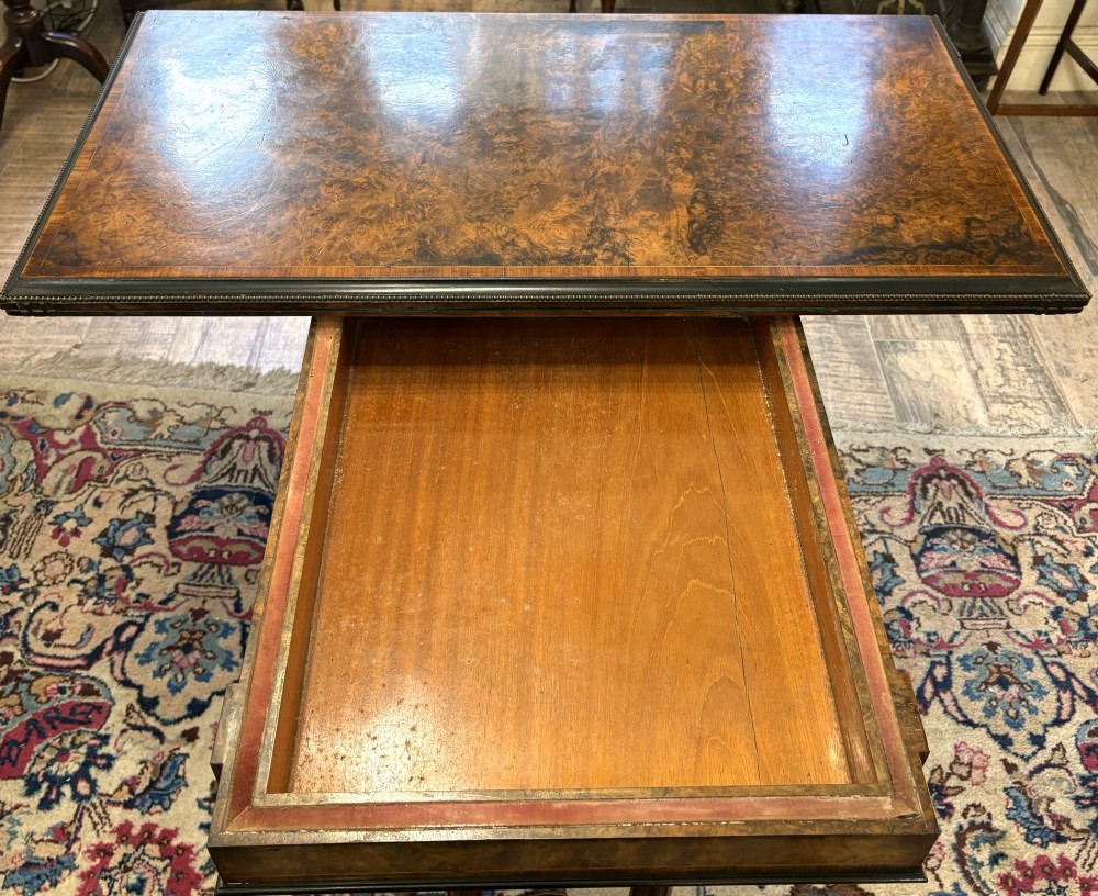 A Victorian burr walnut card table featuring ebonised bead moulding, crossbanded veneer top, twin turned pillar supports, and a turned stretcher base. View IX