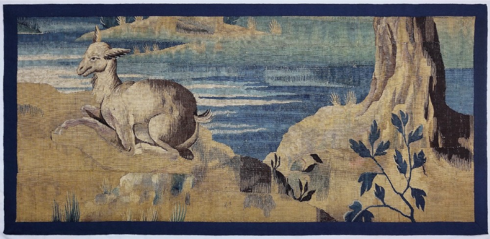 A horizontal 17th-century Flemish woven tapestry panel depicting a horned creature resting in a landscape beside a river and a large tree trunk.