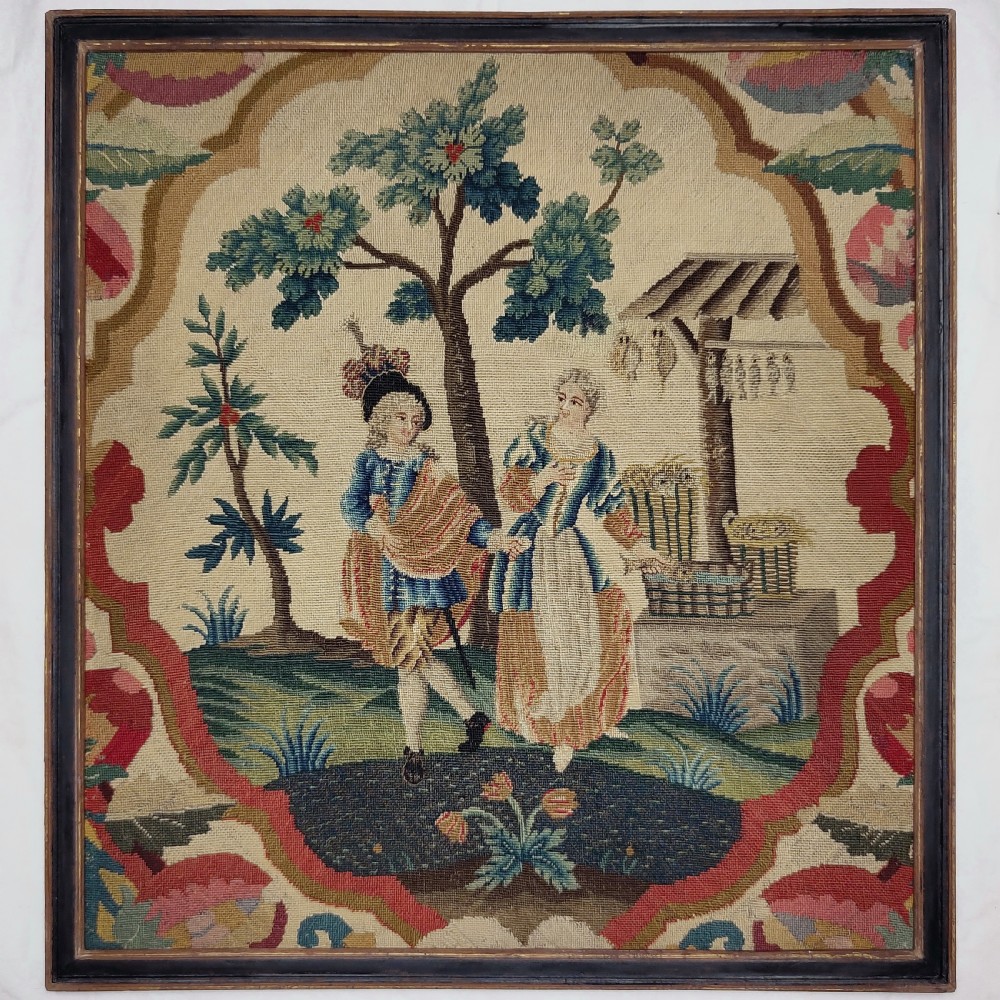 A framed square French needlework panel featuring a pastoral scene of two figures near a well, framed by a red and beige cartouche border.