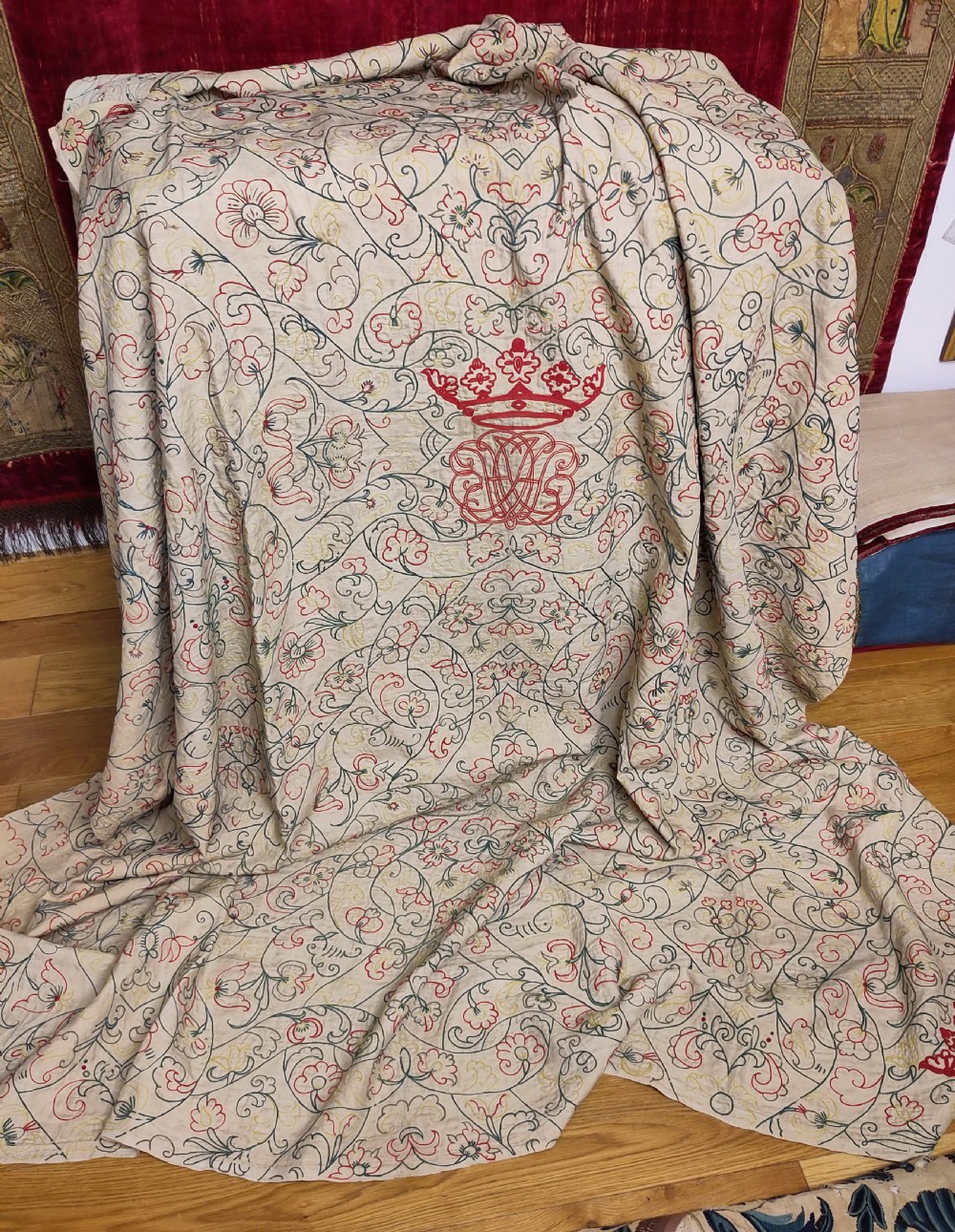 An 18th-century cream linen quilted coverlet featuring intricate floral scrollwork embroidery and a prominent red embroidered coronet and monogram motif at the centre.