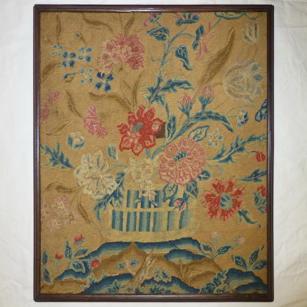An eighteenth-century needlework picture displaying a floral vase arrangement in red, blue, and pink tones on an ochre background inside a slim wooden frame.