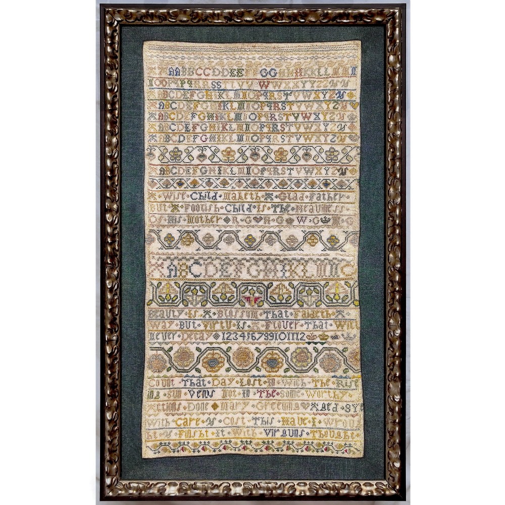 A narrow vertical seventeenth-century framed English band sampler featuring rows of embroidered alphabets, floral bands, and verse on cream linen.