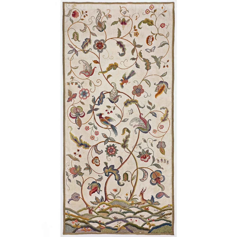 A vertical rectangular crewelwork wall hanging featuring an embroidered Tree of Life motif with scrolling vines, multicoloured flowers, birds, and a deer at the base against a cream background.