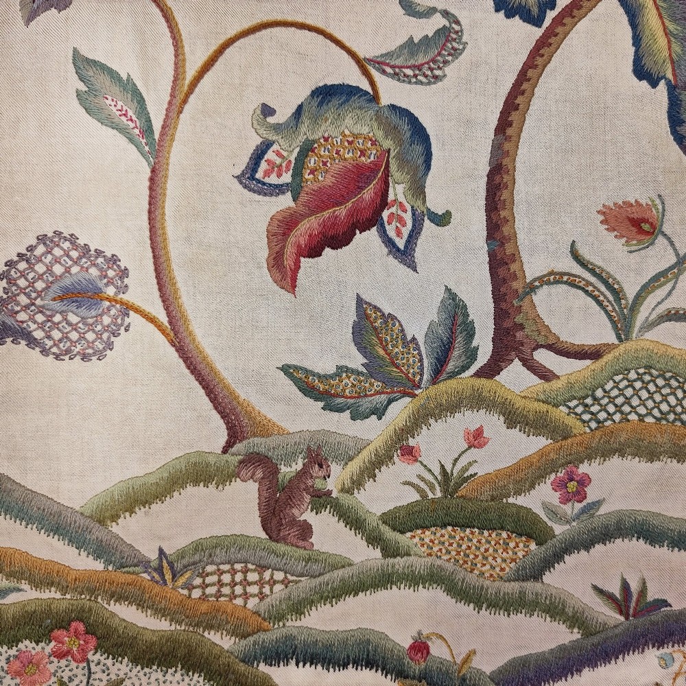 A vertical rectangular crewelwork wall hanging featuring an embroidered Tree of Life motif with scrolling vines, multicoloured flowers, birds, and a deer at the base against a cream background. View IV