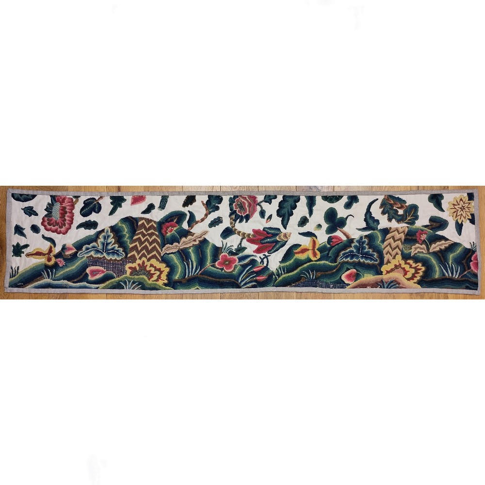 A long horizontal rectangular panel of hand-embroidered English crewelwork featuring stylised green foliage, pink blossoms, and undulating hillocks on a light linen ground.