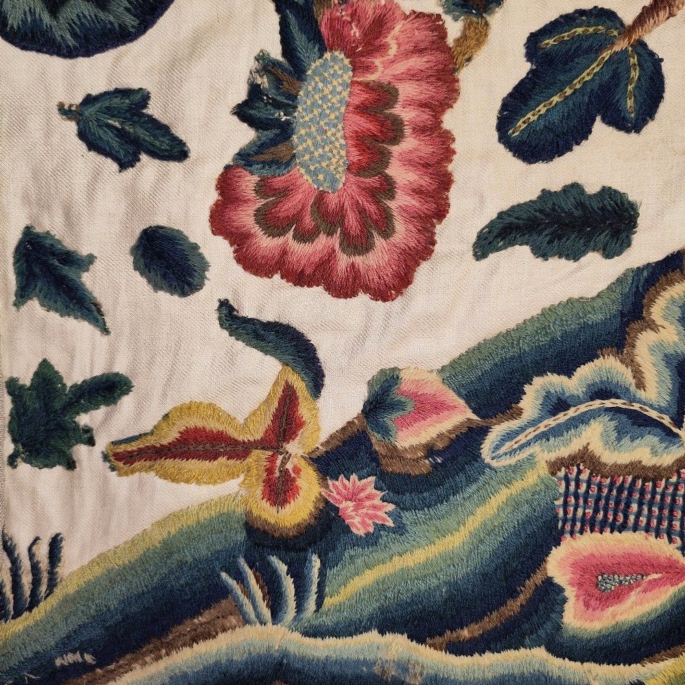 A long horizontal rectangular panel of hand-embroidered English crewelwork featuring stylised green foliage, pink blossoms, and undulating hillocks on a light linen ground. View II