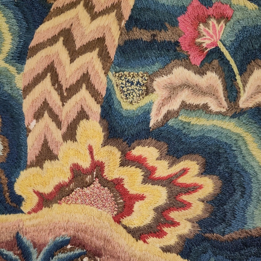 A long horizontal rectangular panel of hand-embroidered English crewelwork featuring stylised green foliage, pink blossoms, and undulating hillocks on a light linen ground. View III