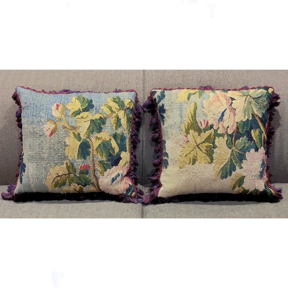 A pair of square cushions constructed from antique Aubusson verdure tapestry featuring botanical foliage in shades of green, yellow, and blue with dark fringed trim.