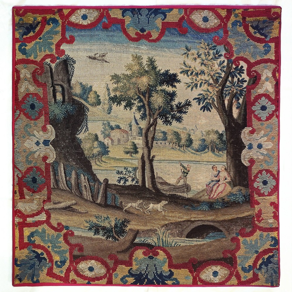 An 18th century square needlework picture depicting a pastoral landscape with figures and dogs framed by an ornamental red and blue strapwork border.