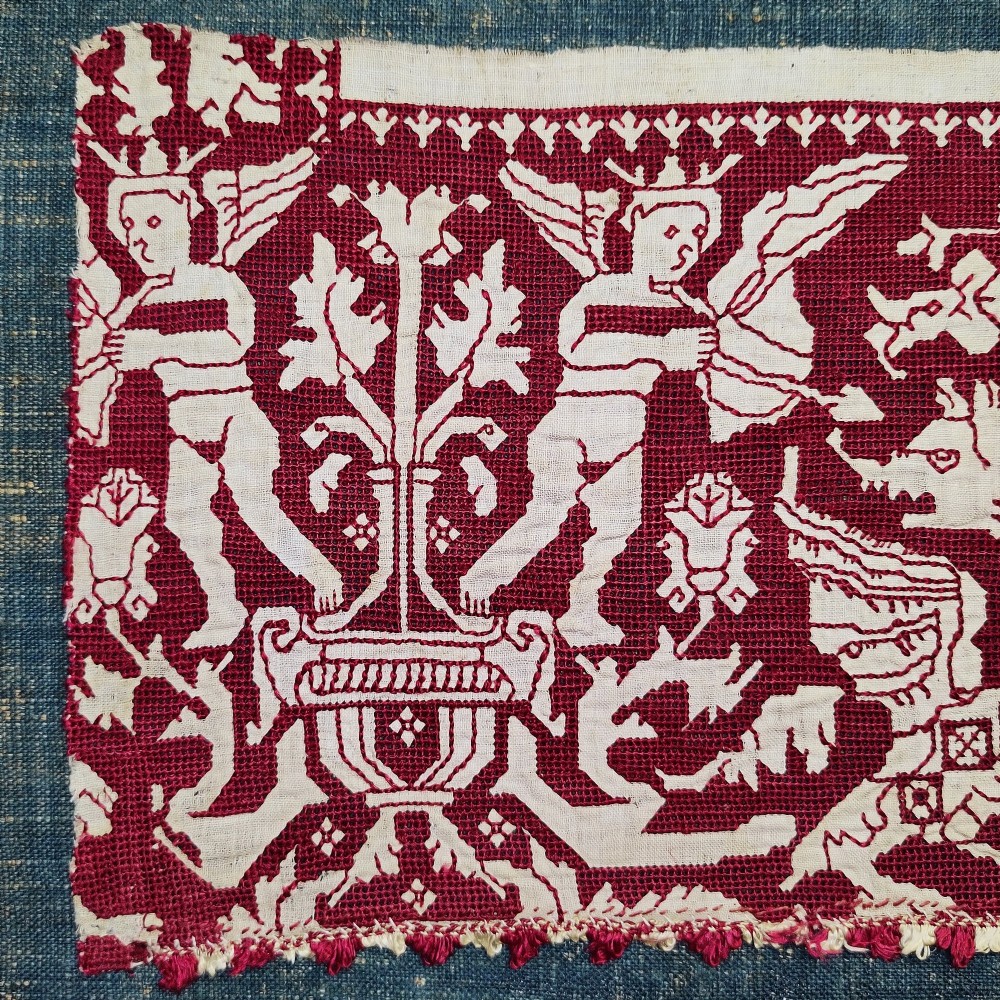 A horizontal 17th-century Italian embroidered red and cream linen border depicting winged figures and mythological creatures, mounted on blue backing within a carved gilt frame. View V