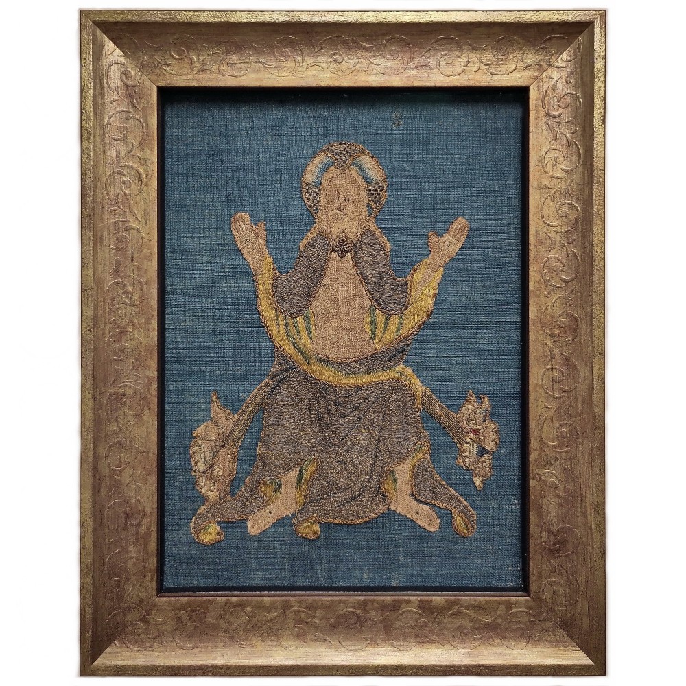 A framed medieval embroidery panel depicting a seated figure with raised hands in metallic thread on a blue fabric backing, enclosed in a carved gold frame.