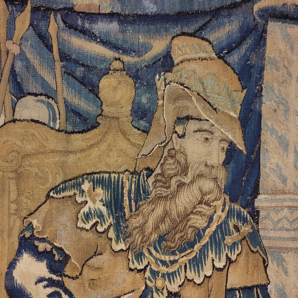 A vertical 16th century Flemish tapestry panel depicting a seated bearded male figure wearing a tall hat and draped robes in blue and gold tones. View II