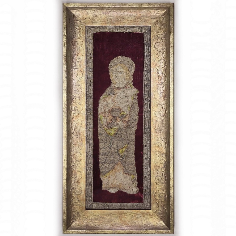 A framed textile art piece featuring an embroidered saint figure mounted on deep burgundy velvet inside a carved gilt wood frame.