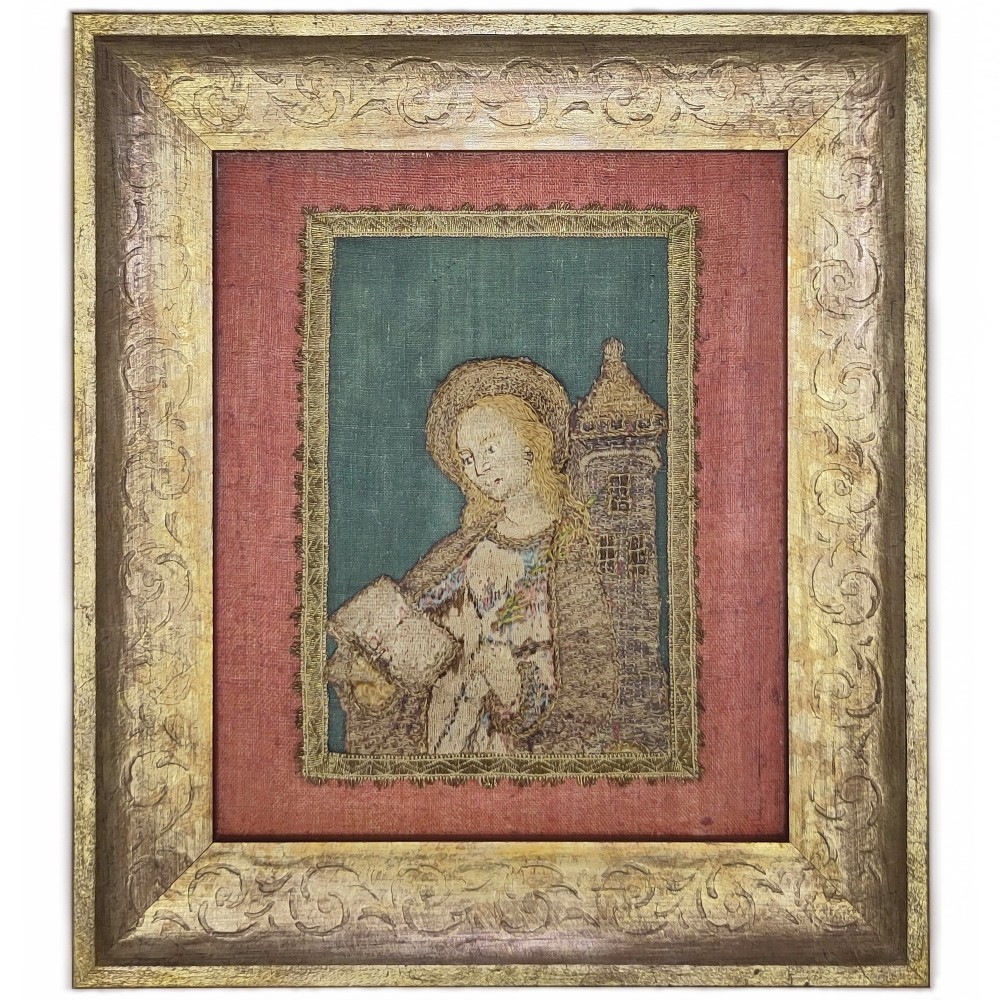 A framed antique embroidered textile fragment depicting a haloed figure holding a book beside a tower, mounted on red fabric within a carved gilt frame.