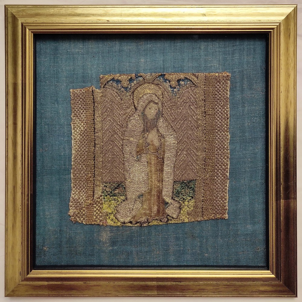 A framed fragment of medieval Opus Anglicanum embroidery featuring a haloed figure embroidered in gold thread, mounted on a blue linen backdrop.