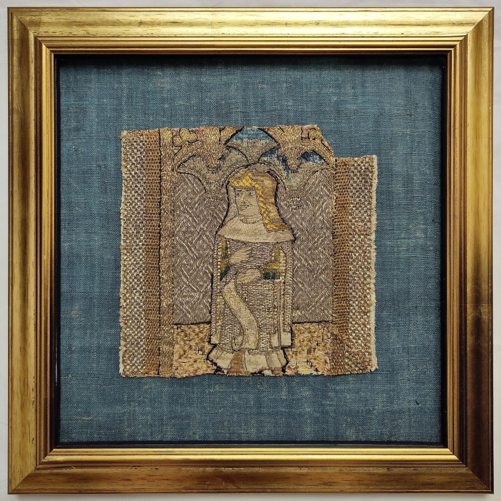 A framed fragment of medieval Opus Anglicanum embroidery depicting a figure beneath a gothic arch, mounted on a blue linen backing within a gilded wooden frame.