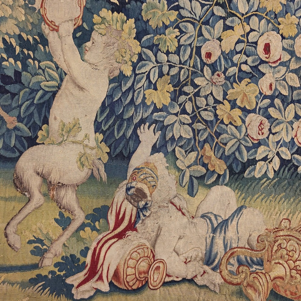 A vertical 17th-century tapestry panel depicting figures and putti amidst dense green trees and foliage. View II