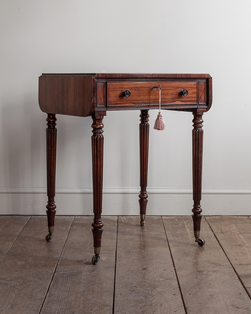 A dark rosewood Regency work table featuring turned fluted legs on brass castors and a single front drawer with a hanging tassel.