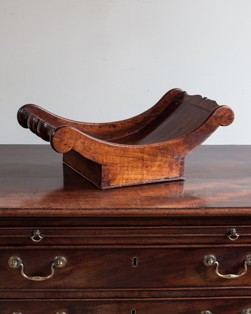 An antique Georgian fruitwood cheese coaster with a crescent body, scrolled terminals, and dovetailed joinery on a rectangular base.