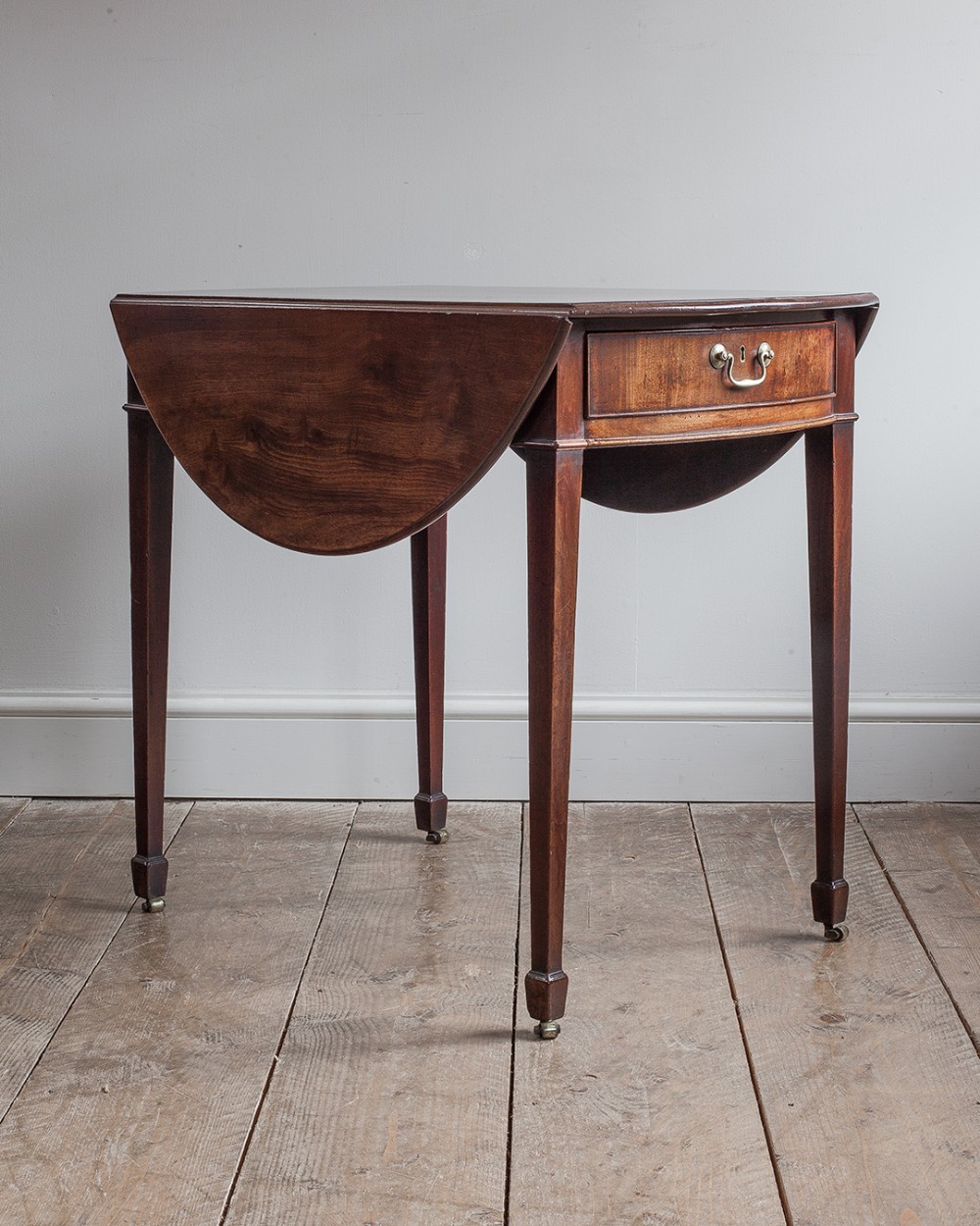 A Georgian mahogany oval Pembroke table featuring two hinged drop leaves, a single frieze drawer with a brass handle, and four square tapered legs on brass castors.