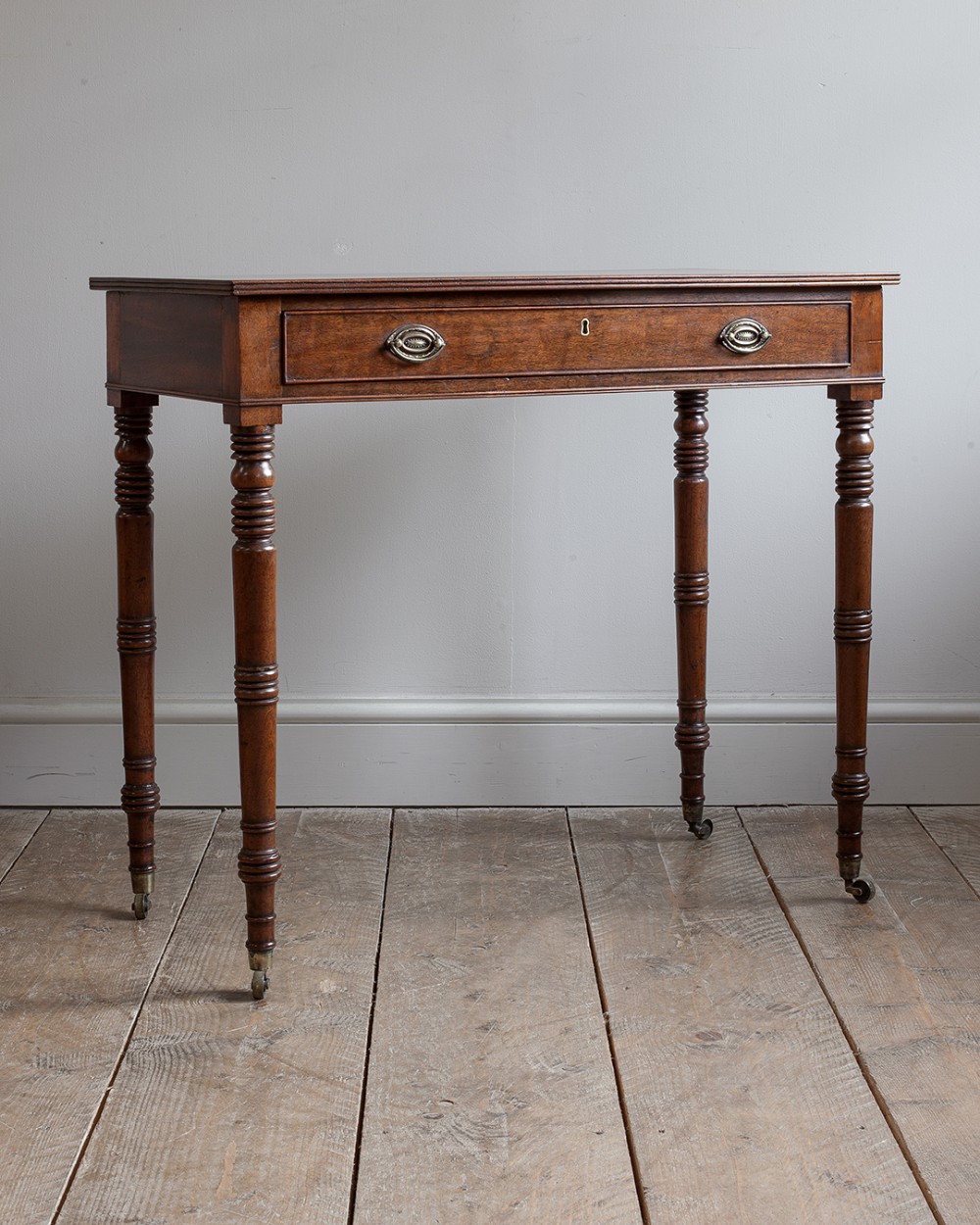A Regency mahogany side table featuring a single frieze drawer with brass oval drop handles, raised on slender turned legs with brass castors.