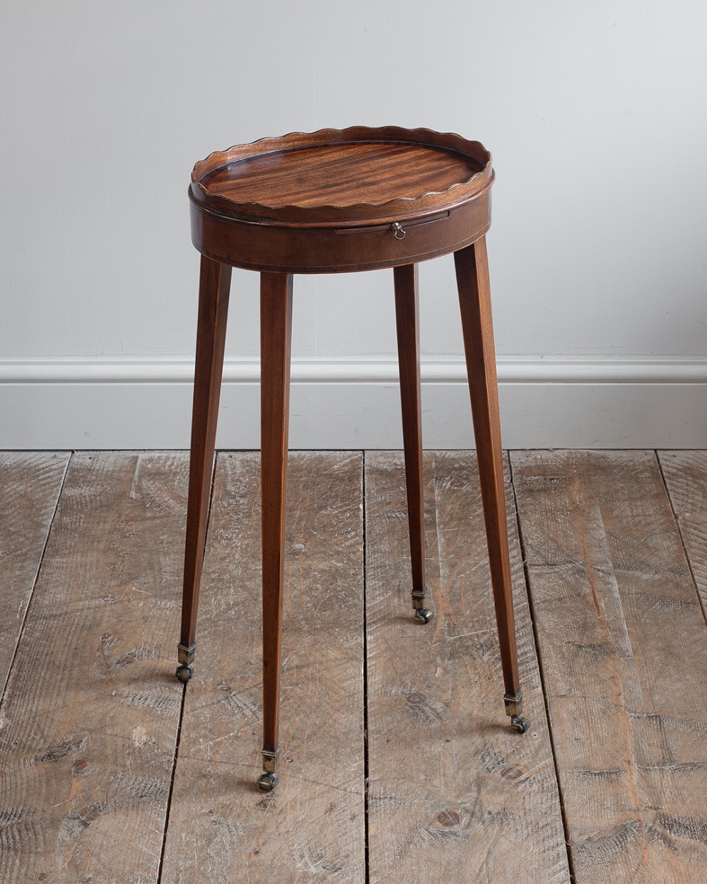 A tall Georgian mahogany oval urn stand featuring a scalloped gallery rim, a pull-out slide, tapered square legs, and brass castors.