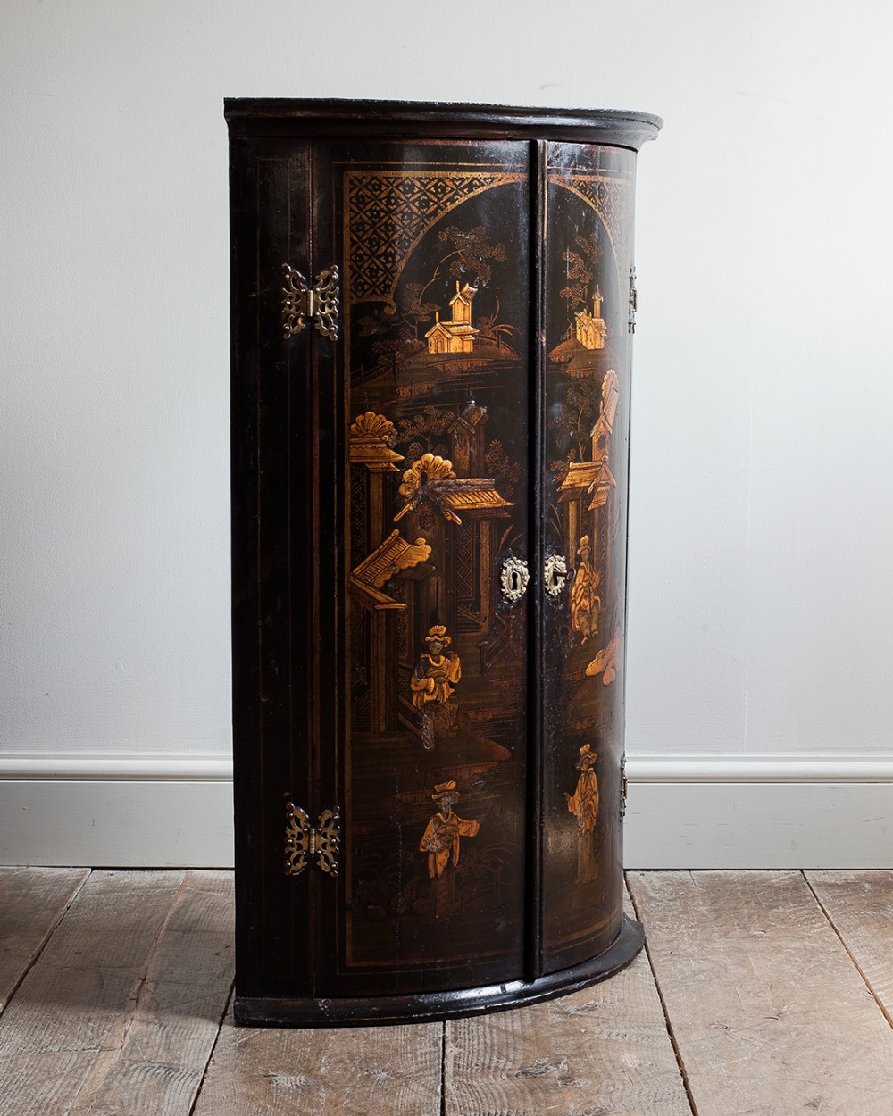 An 18th-century black lacquered bowfront corner cupboard featuring gilt Chinoiserie figures and architecture, fitted with brass butterfly hinges.