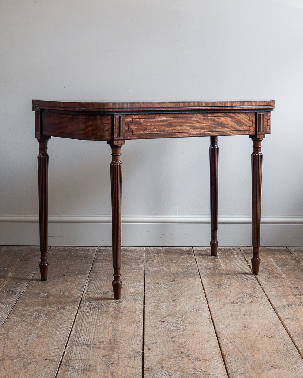 A George IV D-shaped mahogany tea table featuring a fold-over top, frieze with flame veneer inlay, and four reeded tapered legs.