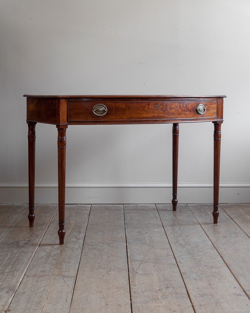 A 19th-century mahogany bowfront side table featuring a single central drawer with brass oval swing handles and four slender turned legs.