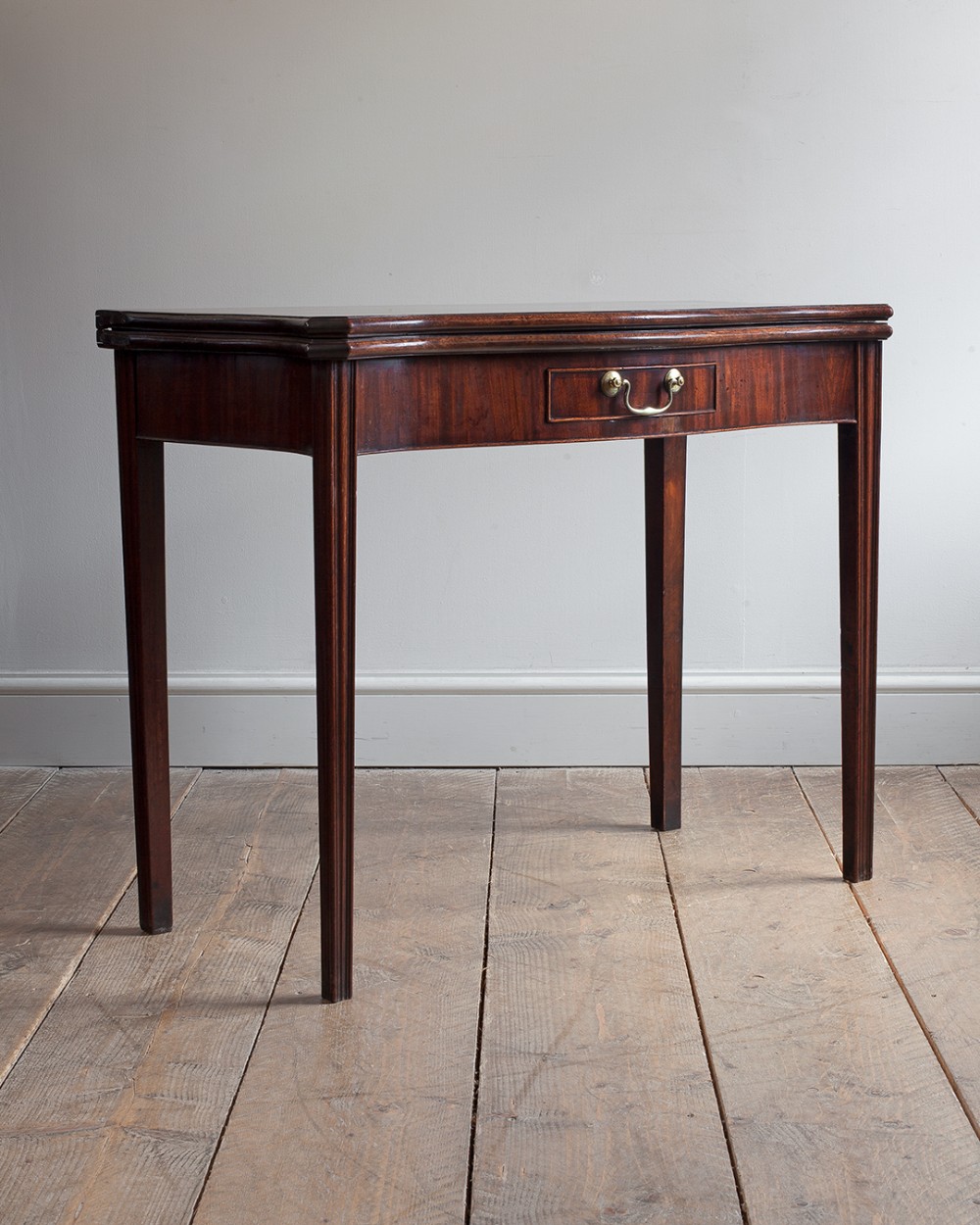 A George III mahogany serpentine card table featuring straight fluted legs, a single central drawer with a brass handle, and a fold-over top.