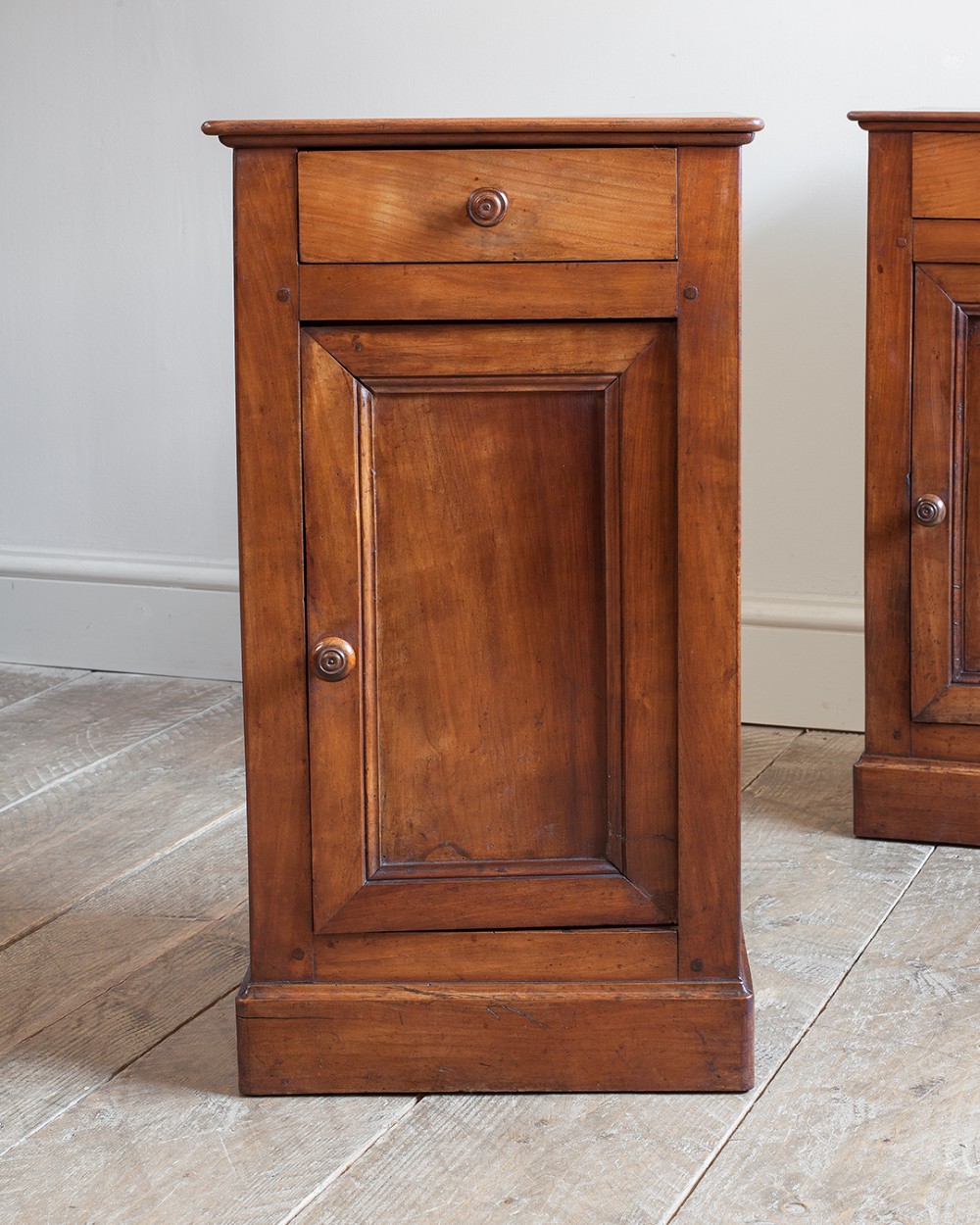 A pair of 19th-century French cherrywood bedside cupboards featuring single top drawers, panelled doors, turned timber knobs, and solid plinth bases. View V