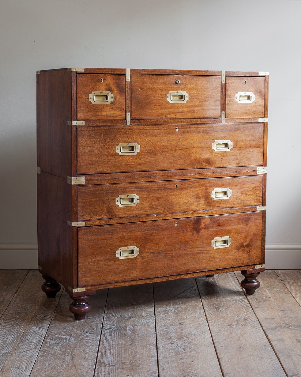 A Victorian two-part teak campaign chest of drawers featuring flush brass handles, protective corner straps, and turned wooden feet.