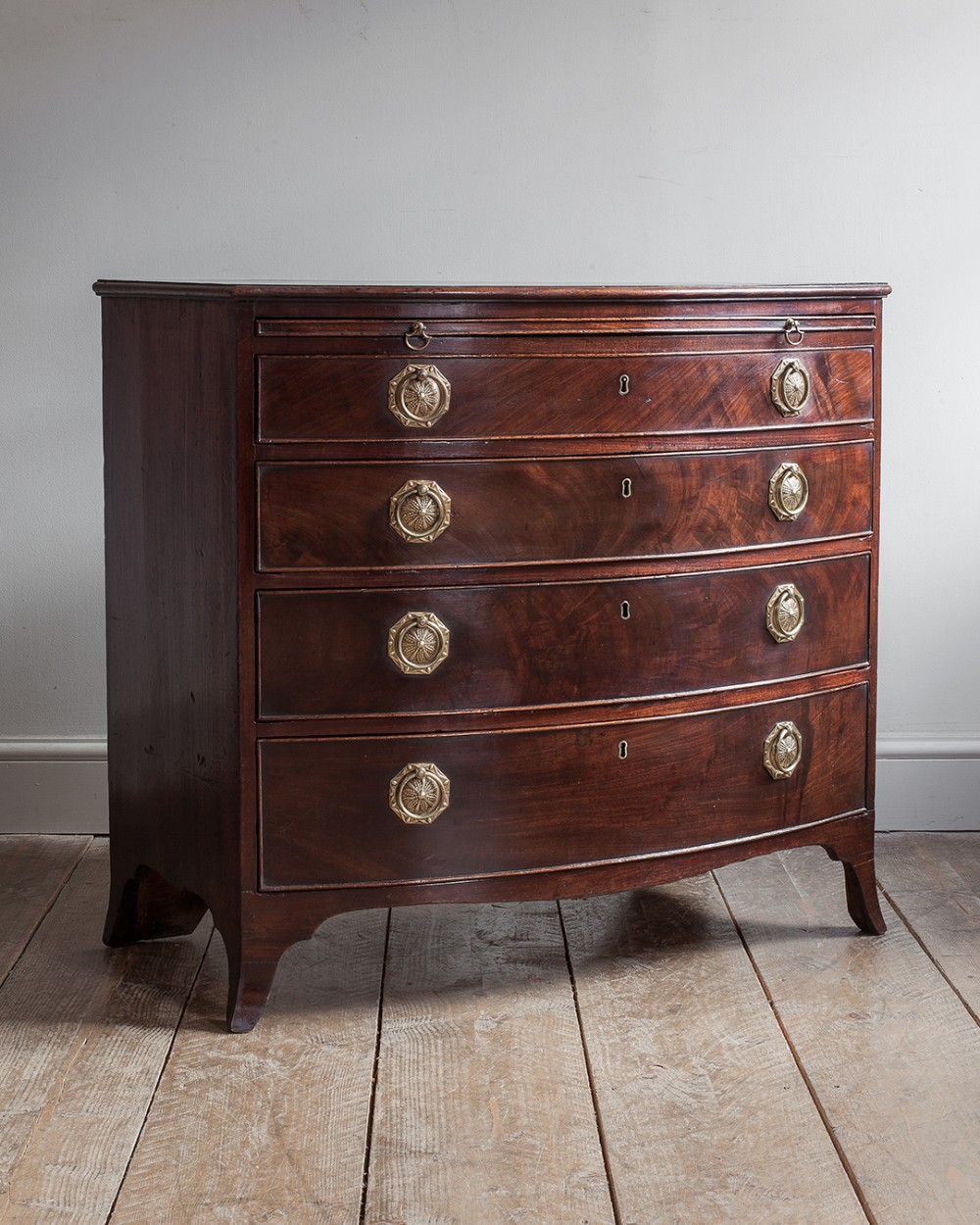 A George III mahogany bowfront chest of drawers featuring four graduated drawers, a pull-out brushing slide, octagonal brass handles, and splayed bracket feet.