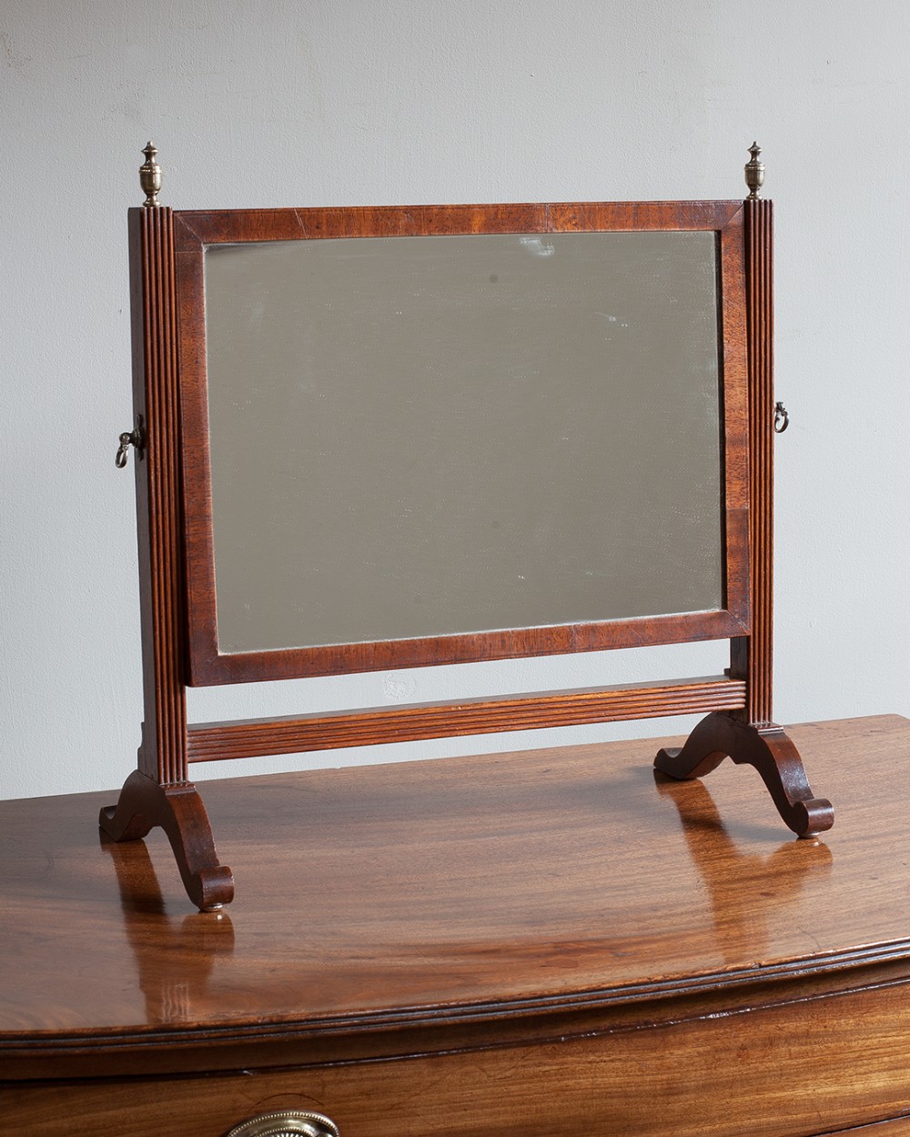 An antique Regency mahogany swing mirror featuring reeded upright columns, brass finials, and outswept sabre feet on a wooden surface.