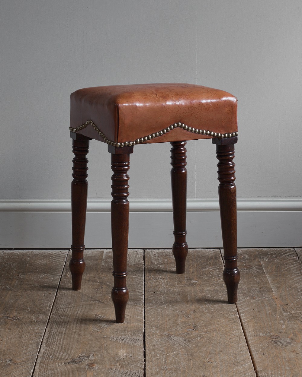 A dark mahogany Regency stool with turned legs and a square top upholstered in tan leather with brass stud detail.